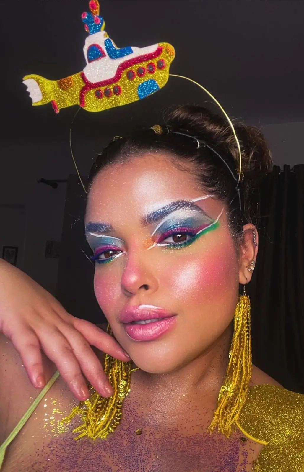 Close-up de uma mulher com maquiagem artística vibrante e colorida, incluindo olhos esfumados com glitter e delineados gráficos, bochechas rosadas e lábios brilhantes. Ela usa uma tiara com um submarino amarelo e brincos longos e dourados.