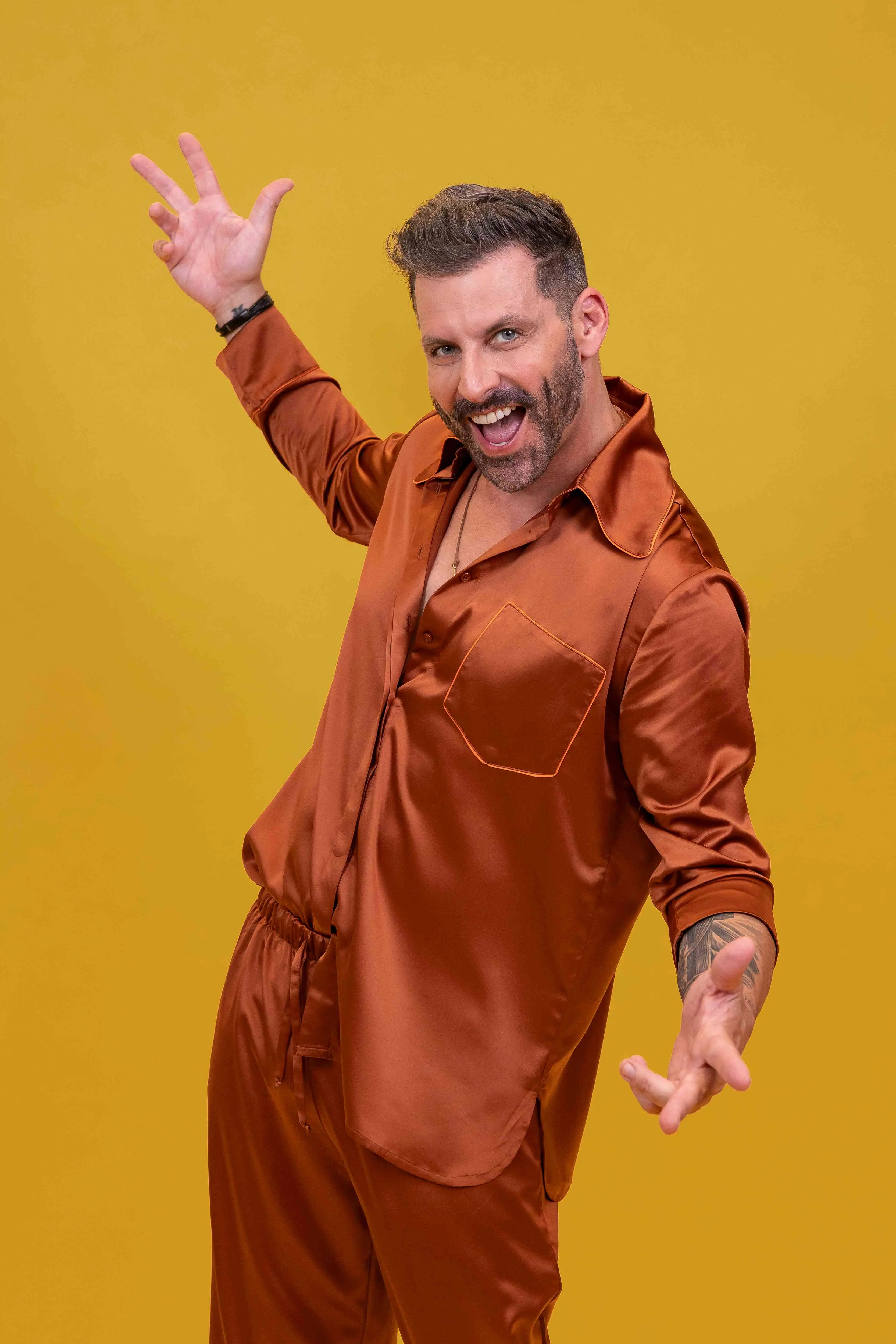Homem com barba e camisa de seda cor bronze, sorrindo e fazendo pose divertida em fundo amarelo monocromático. O homem é Henri Castelli, ator.