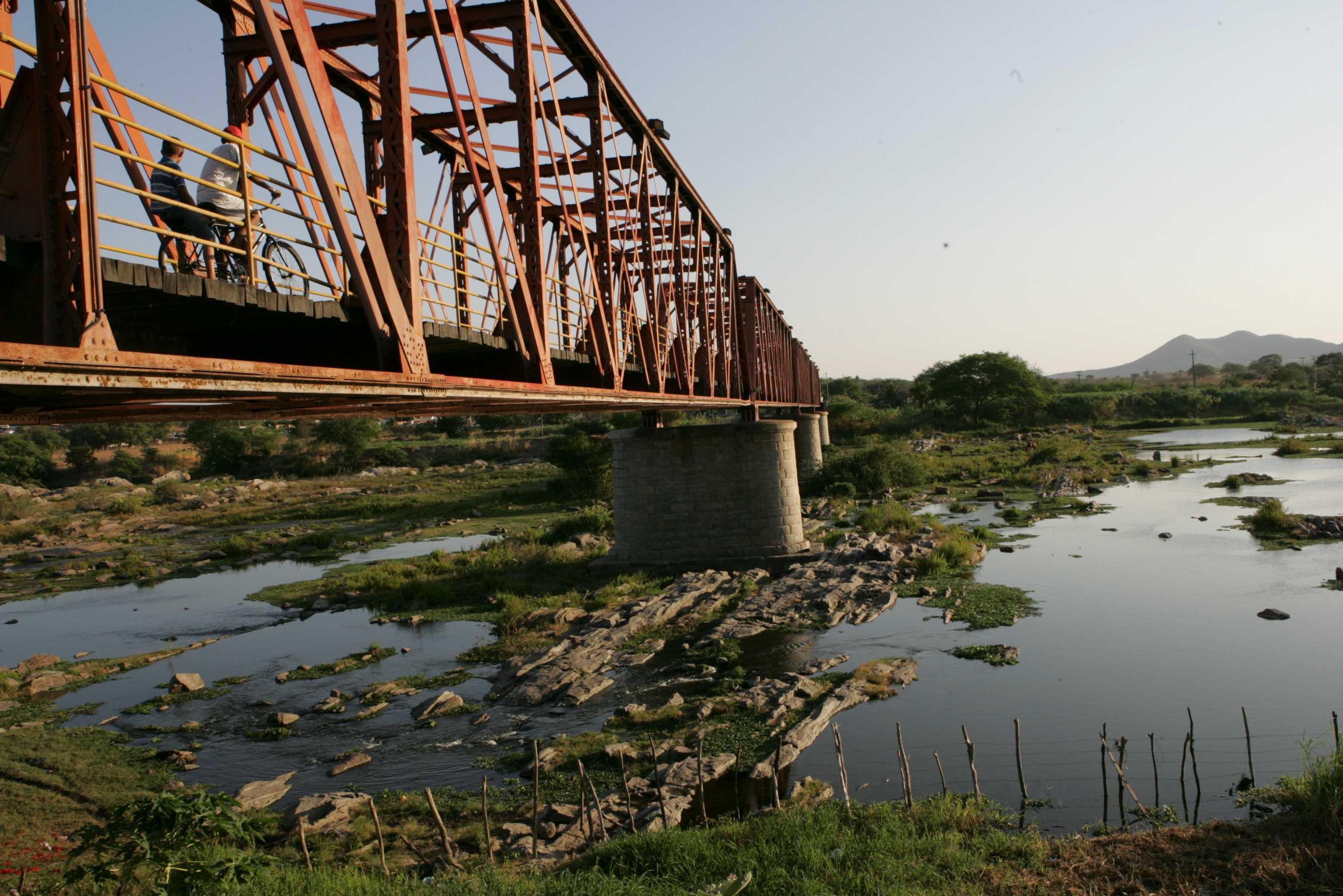 Ponte metálica de Quixeramobim sobre leito rochoso do rio. Pessoas atravessam a estrutura de ferro avermelhada. Ao fundo, vegetação e morros sob céu claro.