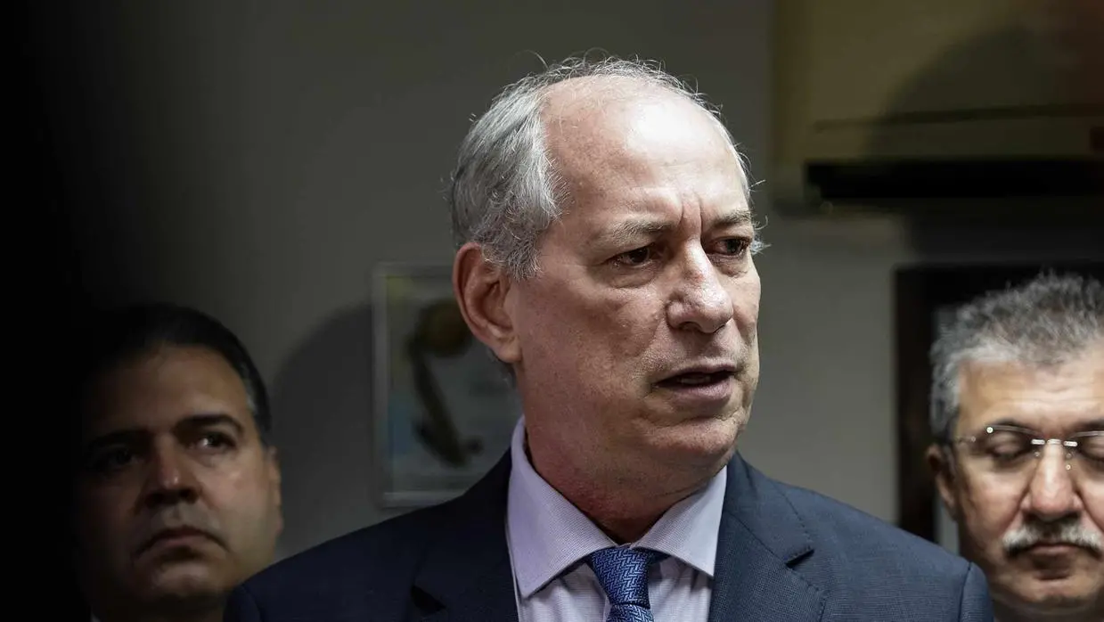 Ciro Gomes vestindo terno e gravata, fala com expressão séria em ambiente interno. Outras duas pessoas aparecem desfocadas atrás dele, acompanhando a cena.