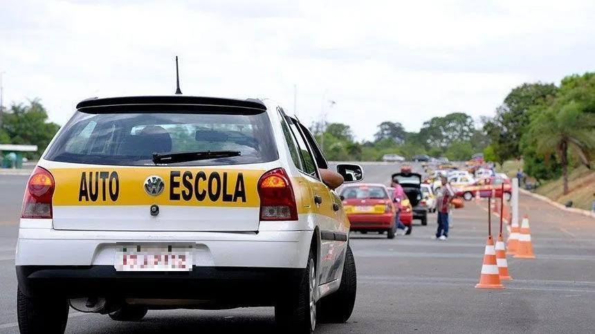 Carro de autoescola visto por trás, parado em pista de prova prática de direção, com cones laranja organizando o percurso e outros veículos ao fundo durante exame de habilitação.