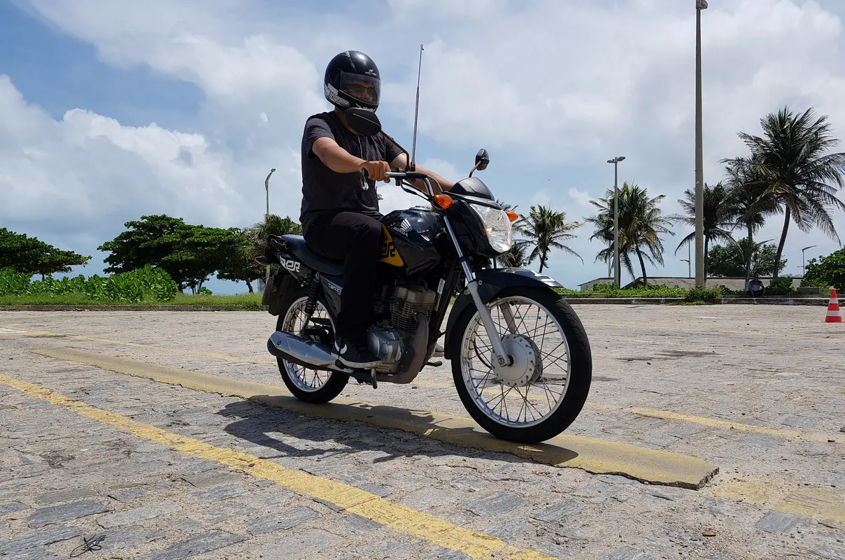 Homem em treinamento de pilotagem de moto, mantendo o equilíbrio em uma motocicleta preta sobre um obstáculo de linha elevada amarela, com cones e árvores no entorno.