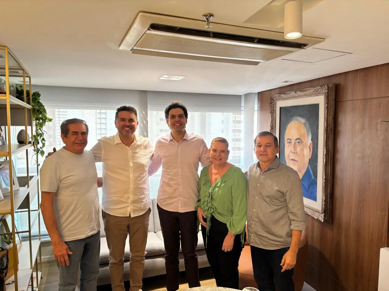 Cinco pessoas posam lado a lado em uma sala de estar iluminada, com sofá ao fundo, estante com objetos decorativos à esquerda e um retrato emoldurado na parede à direita. Elas vestem roupas casuais e sociais claras e sorriem para a câmera.