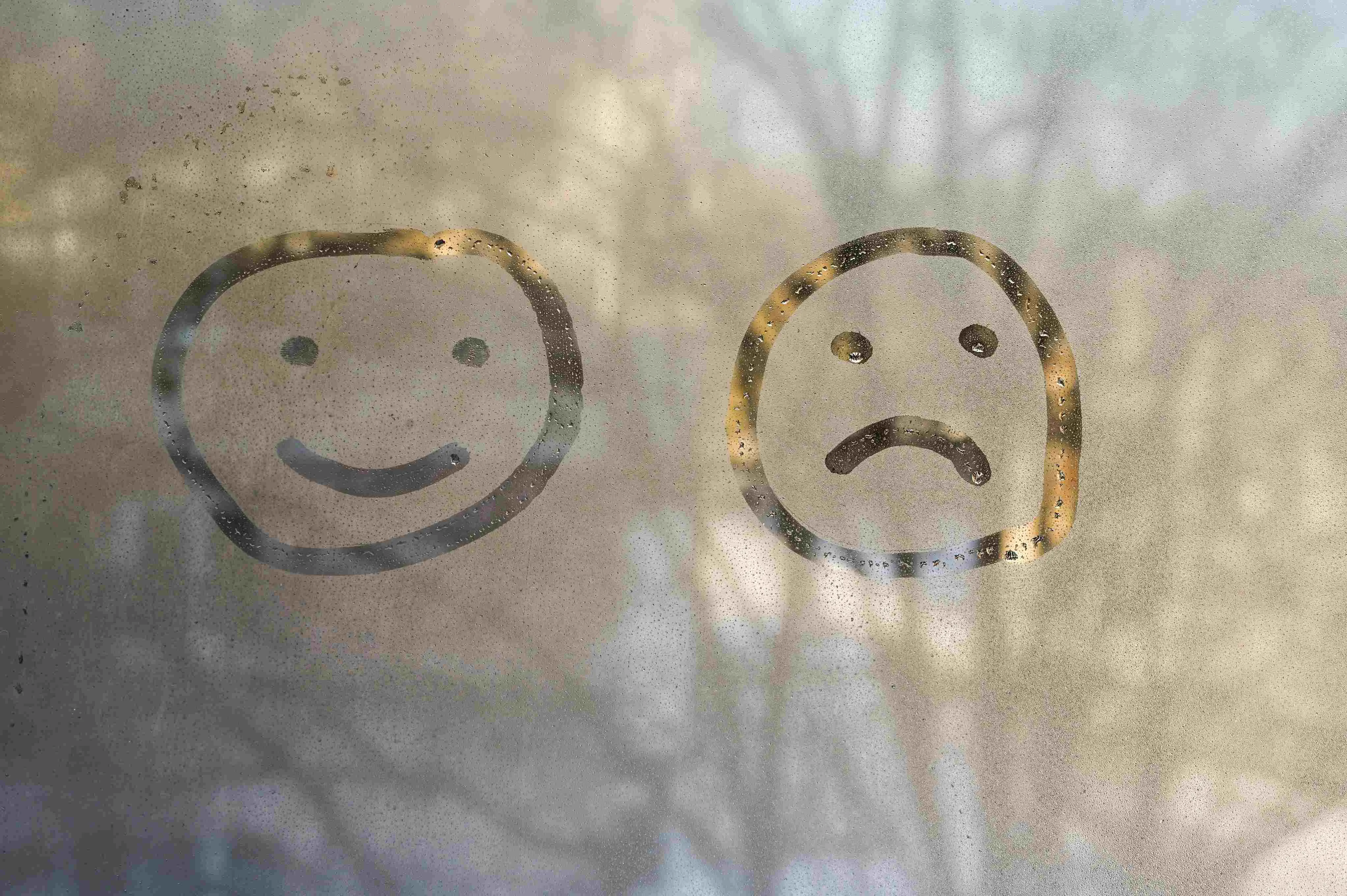 Desenhos de um rosto feliz e um rosto triste feitos no vapor de um vidro embaçado. Ao fundo, a silhueta de árvores aparece desfocada pela condensação e pela luz externa.