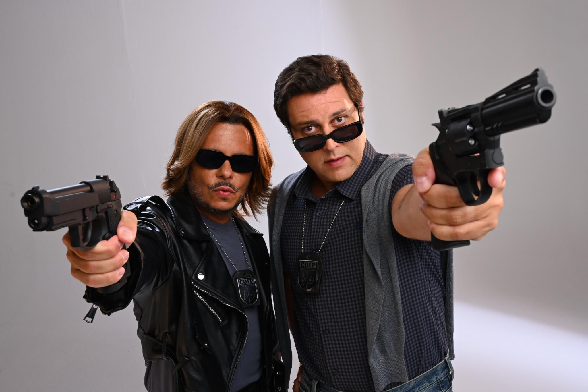 Dois agentes seguram pistolas apontadas para a câmera. Ambos usam roupas escuras e distintivos pendurados no pescoço, posando lado a lado em um estúdio de fundo neutro em uma postura firme e alerta.