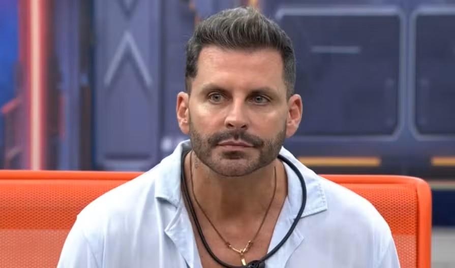 Homem com cabelo escuro e barba, vestindo camisa azul clara e colares, com um olhar direto e sério. Ele está sentado em uma cadeira laranja, com um fundo desfocado que sugere um cenário de estúdio.