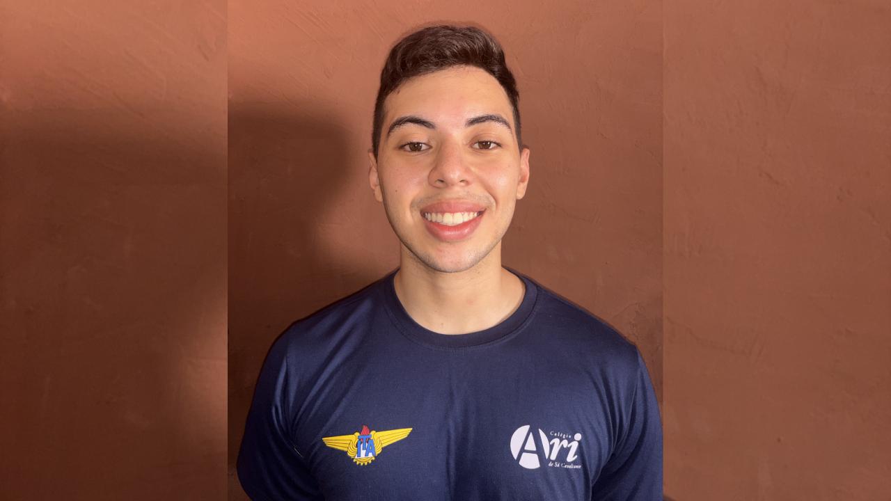 Retrato de um jovem sorrindo, usando uma camiseta azul-marinho com os logotipos do Colégio Ari de Sá Cavalcante e um emblema dourado de asas no peito, do ITA.