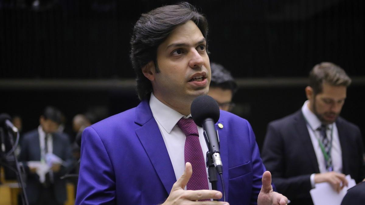 AJ Albuquerque de terno azul e gravata roxa fala ao microfone em um plenário, gesticulando com as mãos; ao fundo, outras pessoas de terno aparecem desfocadas, algumas lendo documentos.