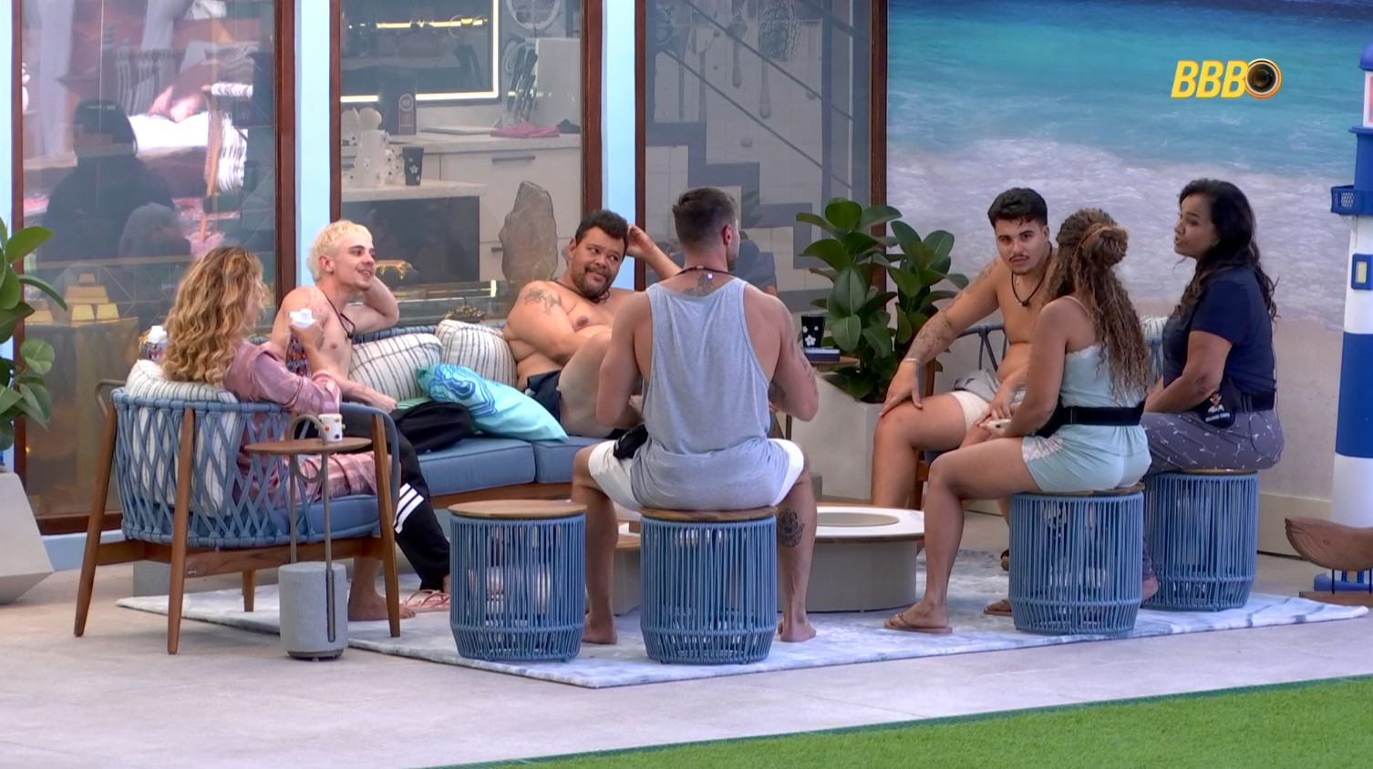 Um grupo de sete participantes do Big Brother Brasil (BBB) conversando em uma área externa da casa, com um grande mural de praia ao fundo e o logotipo do BBB visível.