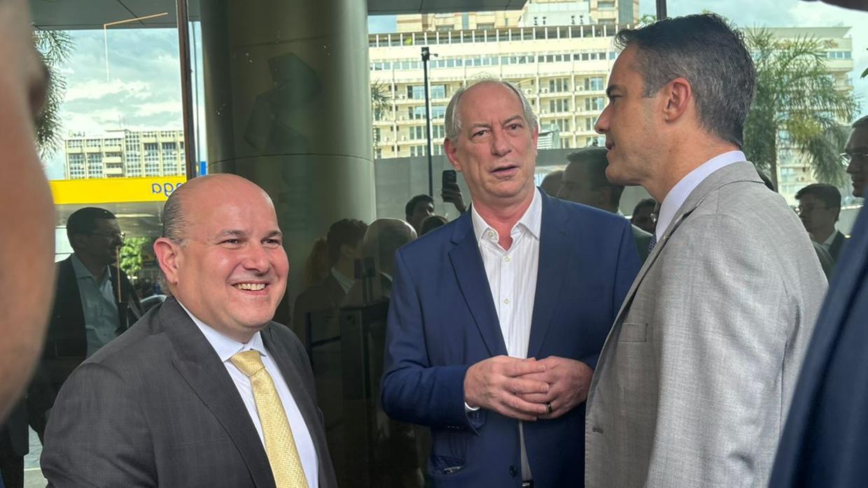 Roberto Cláudio, Ciro Gomes e Capitão Wagner em trajes formais conversam sorrindo na entrada de um prédio envidraçado. A cena mostra um ambiente de evento político ou institucional, com outros participantes ao fundo e um dia ensolarado refletido nas janelas.