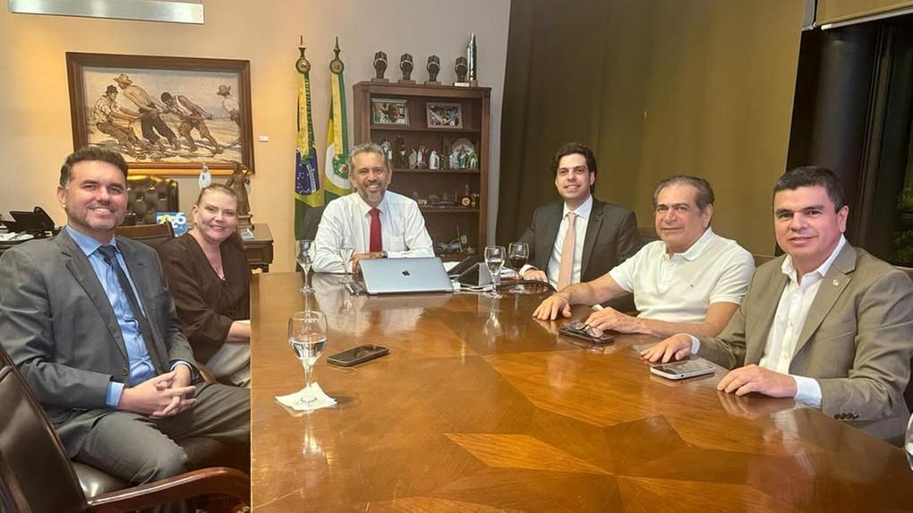 Sete pessoas sentadas ao redor de uma grande mesa de madeira em uma sala de reuniões formal. Elas vestem roupas sociais e estão sorrindo para a câmera. Sobre a mesa há copos de água, celulares e um notebook. Ao fundo, vê-se um escritório com estantes, quadros, objetos decorativos e bandeiras do Brasil e de um estado.