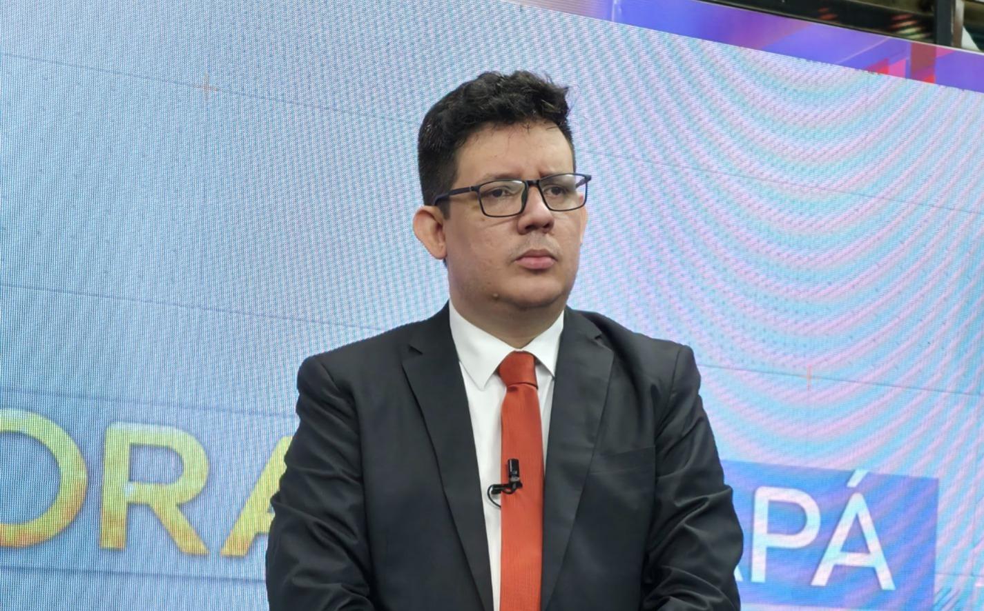 Homem de óculos, usando terno preto, camisa branca e gravata vermelha, em pé diante de um painel luminoso de estúdio. Ele tem expressão séria e usa um microfone preso à lapela.