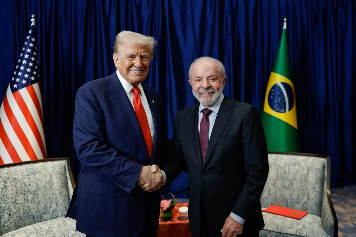 Trump e Lula, de terno, trocam cumprimentos. Ao fundo, a bandeira dos ERUA e do Brasil.