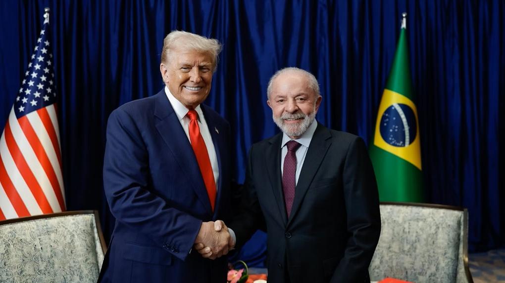 Trump e Lula, de terno, trocam cumprimentos. Ao fundo, a bandeira dos ERUA e do Brasil.