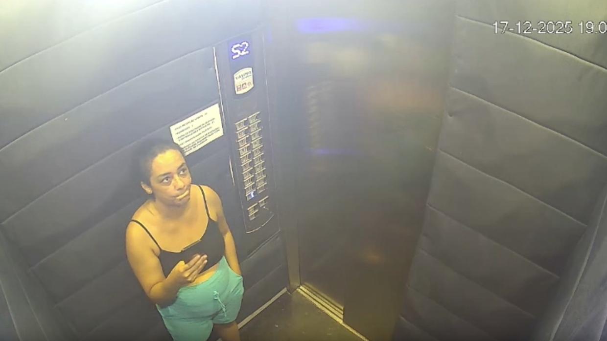 Imagem de câmera de segurança dentro de um elevador: mulher de cabelos presos, usando blusa preta de alça e short verde, olha para cima e faz um gesto com a mão. O painel de botões do elevador aparece ao fundo e a imagem tem aparência granulada.