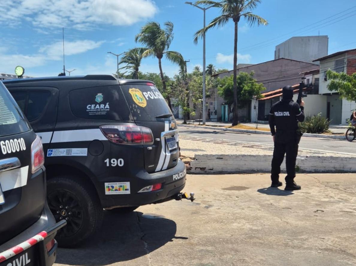 Viaturas da Polícia Civil do Ceará estacionadas em via urbana; ao fundo, um agente de costas, com uniforme preto, aponta uma arma longa em direção a uma rua residencial com casas e coqueiros sob céu azul.