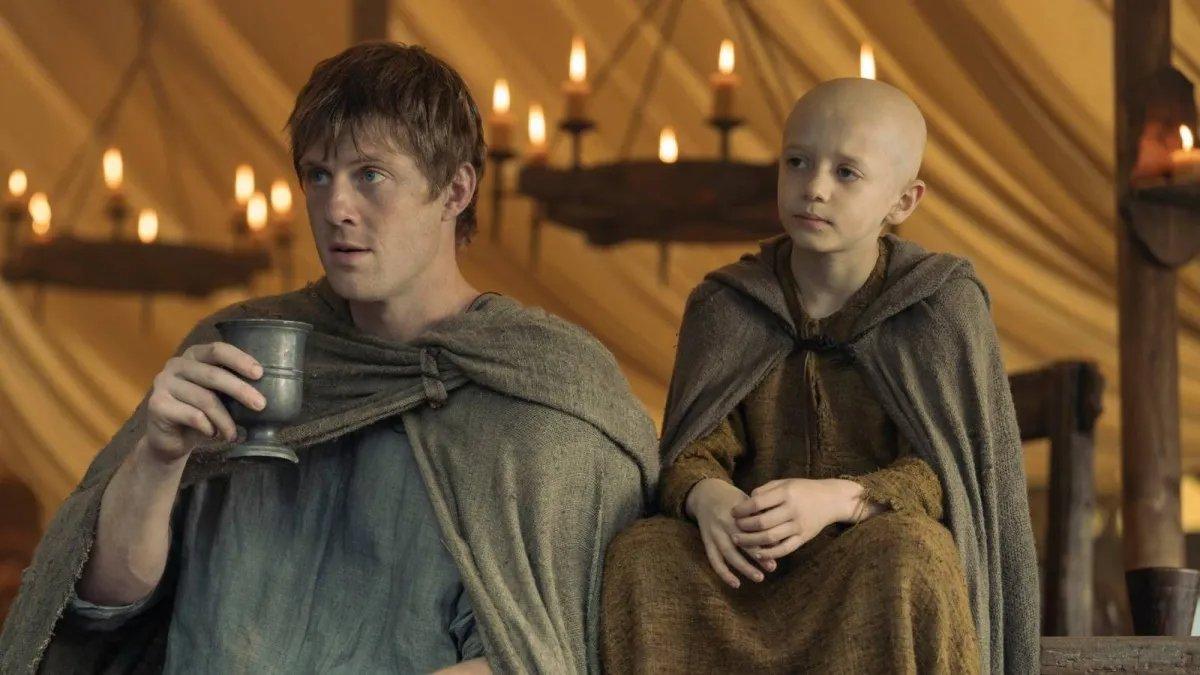 Cena da série “O Cavaleiro dos Sete Reinos” mostra dois personagens com mantos simples em ambiente medieval iluminado por velas.