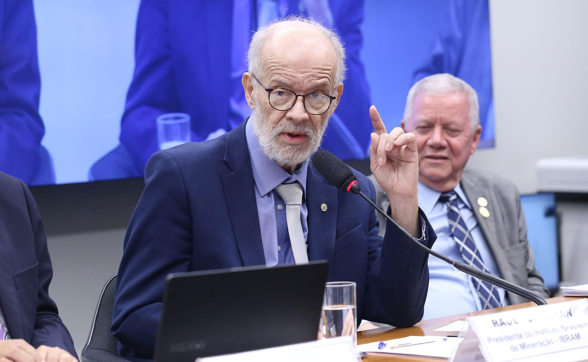 Raul Jungmann com terno e gravata, com o dedo indicador levantado, falando em um evento formal, com uma tela e um segundo indivíduo visíveis ao fundo.