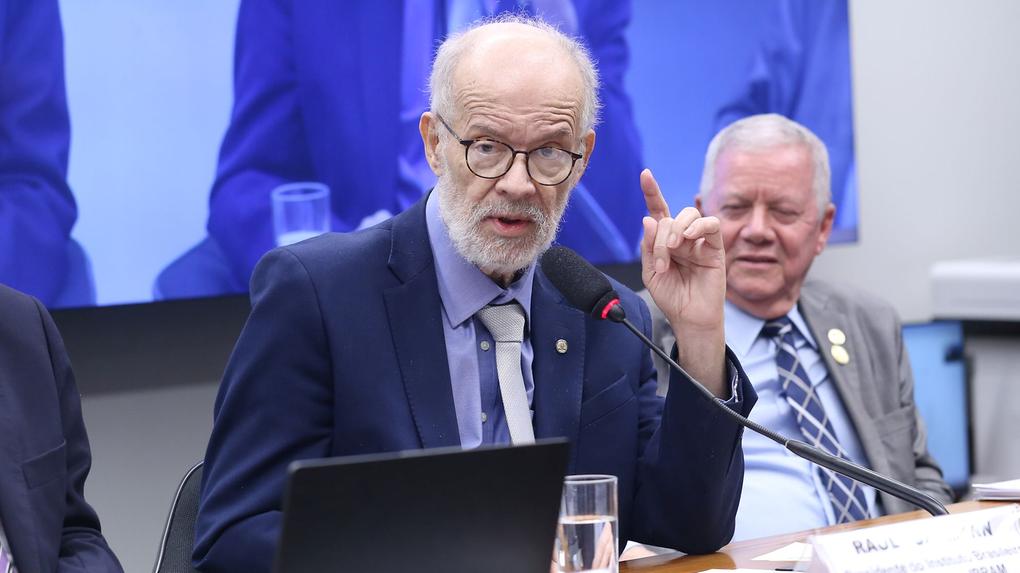 Raul Jungmann com terno e gravata, com o dedo indicador levantado, falando em um evento formal, com uma tela e um segundo indivíduo visíveis ao fundo.