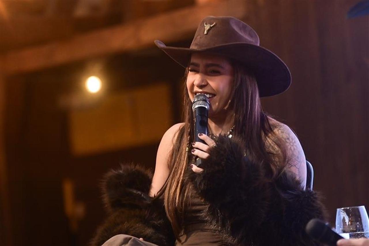 Mulher branca de cabelos castanhos usando um chapéu de cowboy marrom, regata marrom e casaco preto com pelos caindo pelos ombros. Ela segura um microfone e sorri.