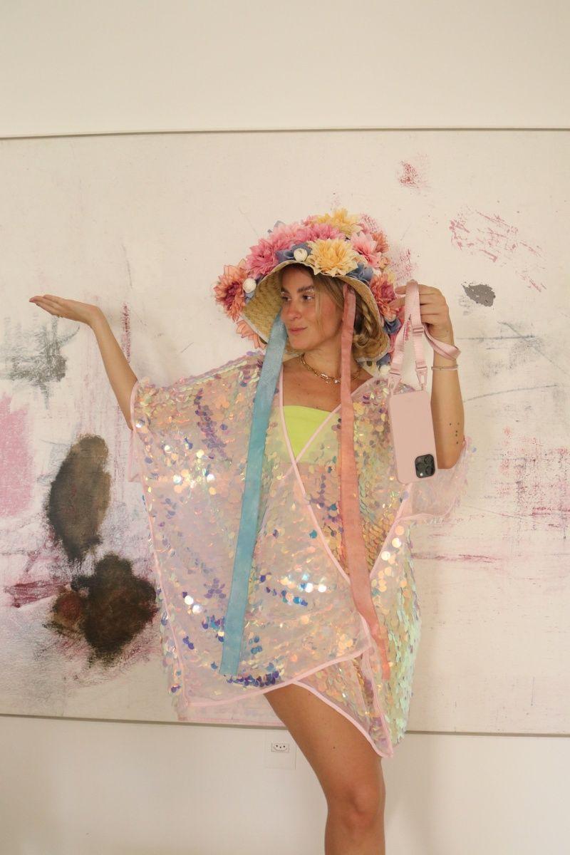 Mulher com chapéu extravagante decorado com flores coloridas e caftan de lantejoulas brilhantes, posando em frente a uma obra de arte abstrata.