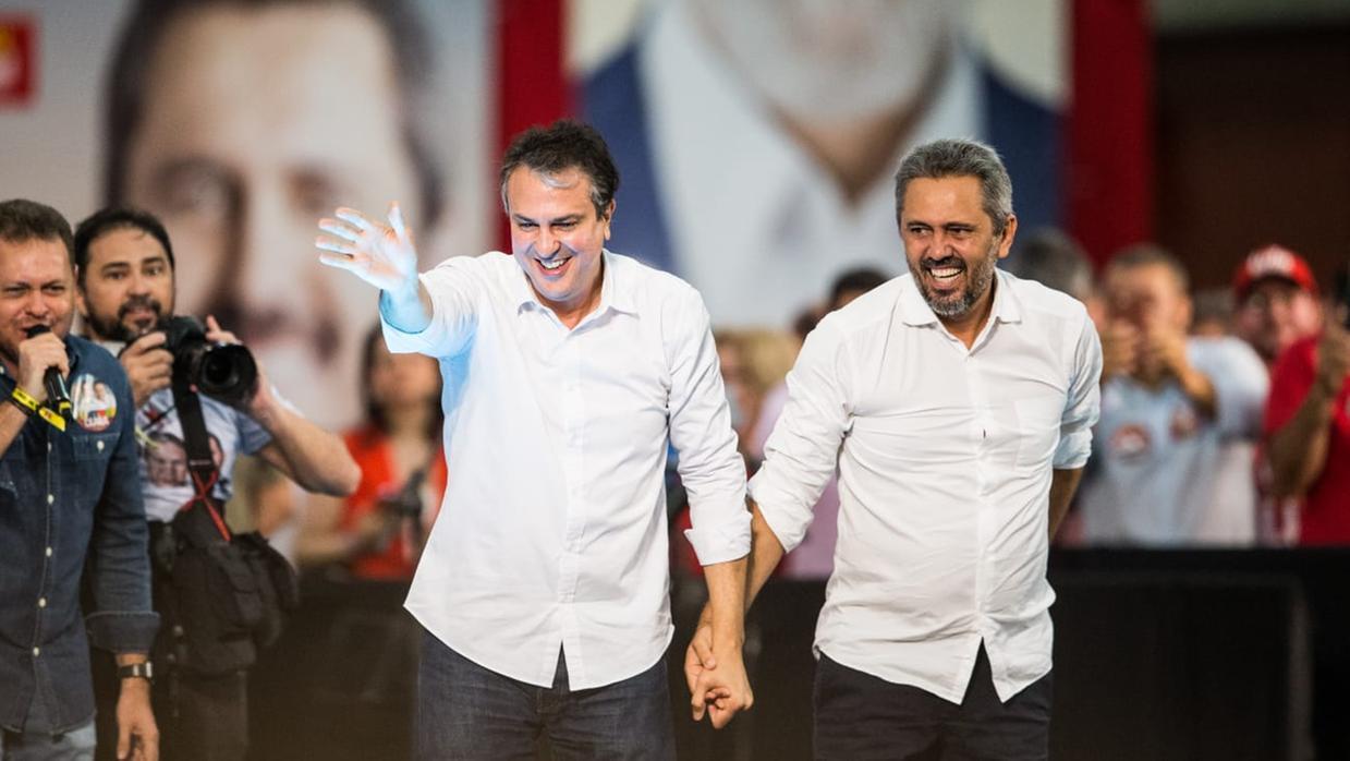 Dois políticos de camisa branca, Camilo e Elmano,  caminham de mãos dadas sobre um palco, sorrindo e acenando para o público durante um ato político; ao fundo, apoiadores e um grande painel com os rostos de Elmano e Lula.