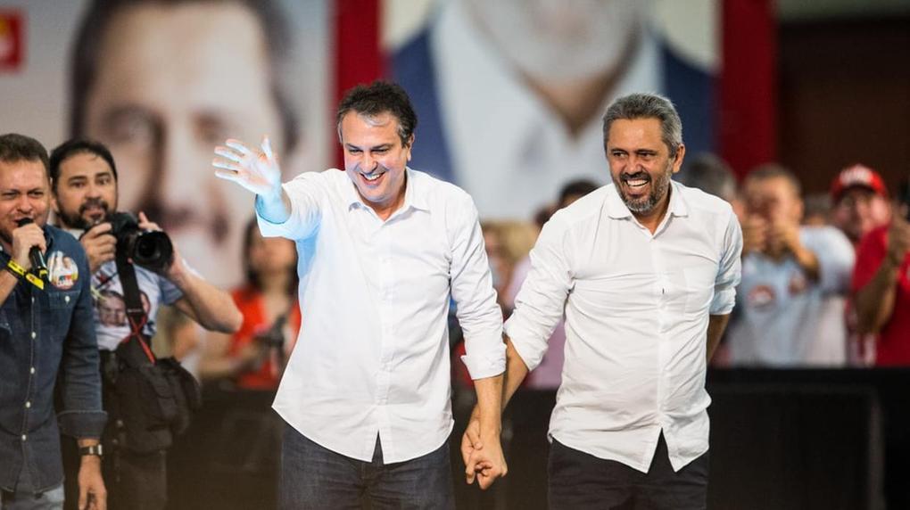 Dois políticos de camisa branca, Camilo e Elmano, caminham de mãos dadas sobre um palco, sorrindo e acenando para o público durante um ato político; ao fundo, apoiadores e um grande painel com os rostos de Elmano e Lula.