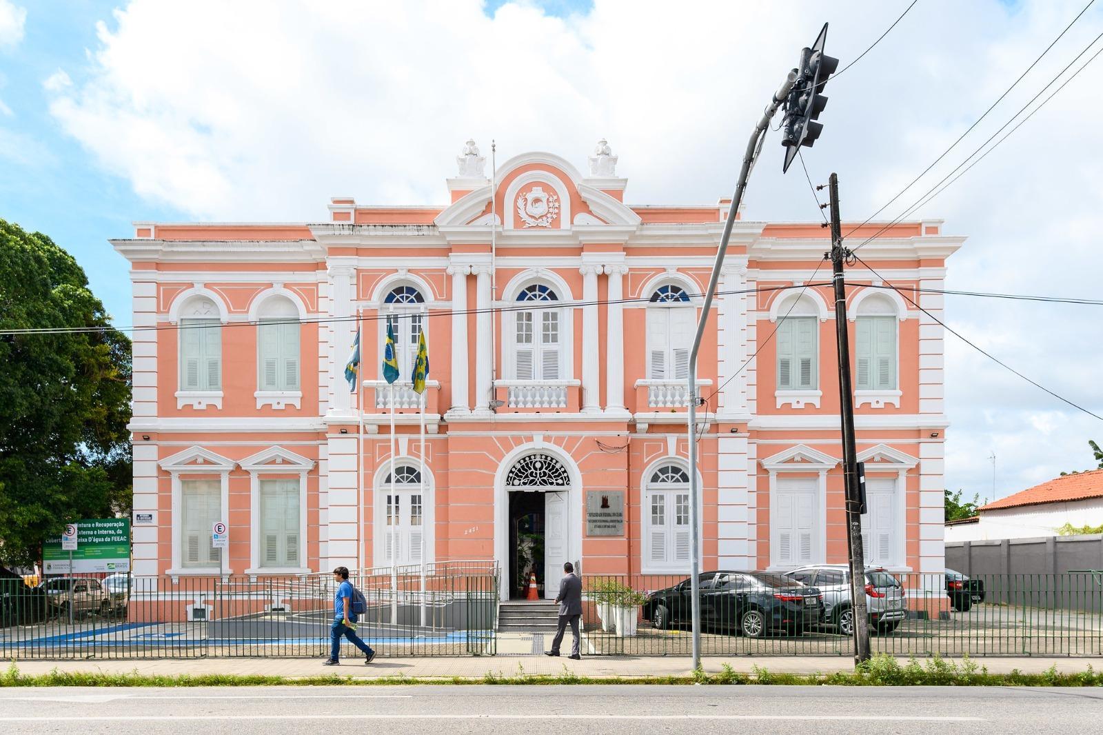 Prédio da UFC na Avenida da Universidade com arquitetura clássica em cor-de-rosa claro e janelas brancas, com a bandeira do Brasil e outra local visíveis em sua fachada elaborada, em um cenário urbano.