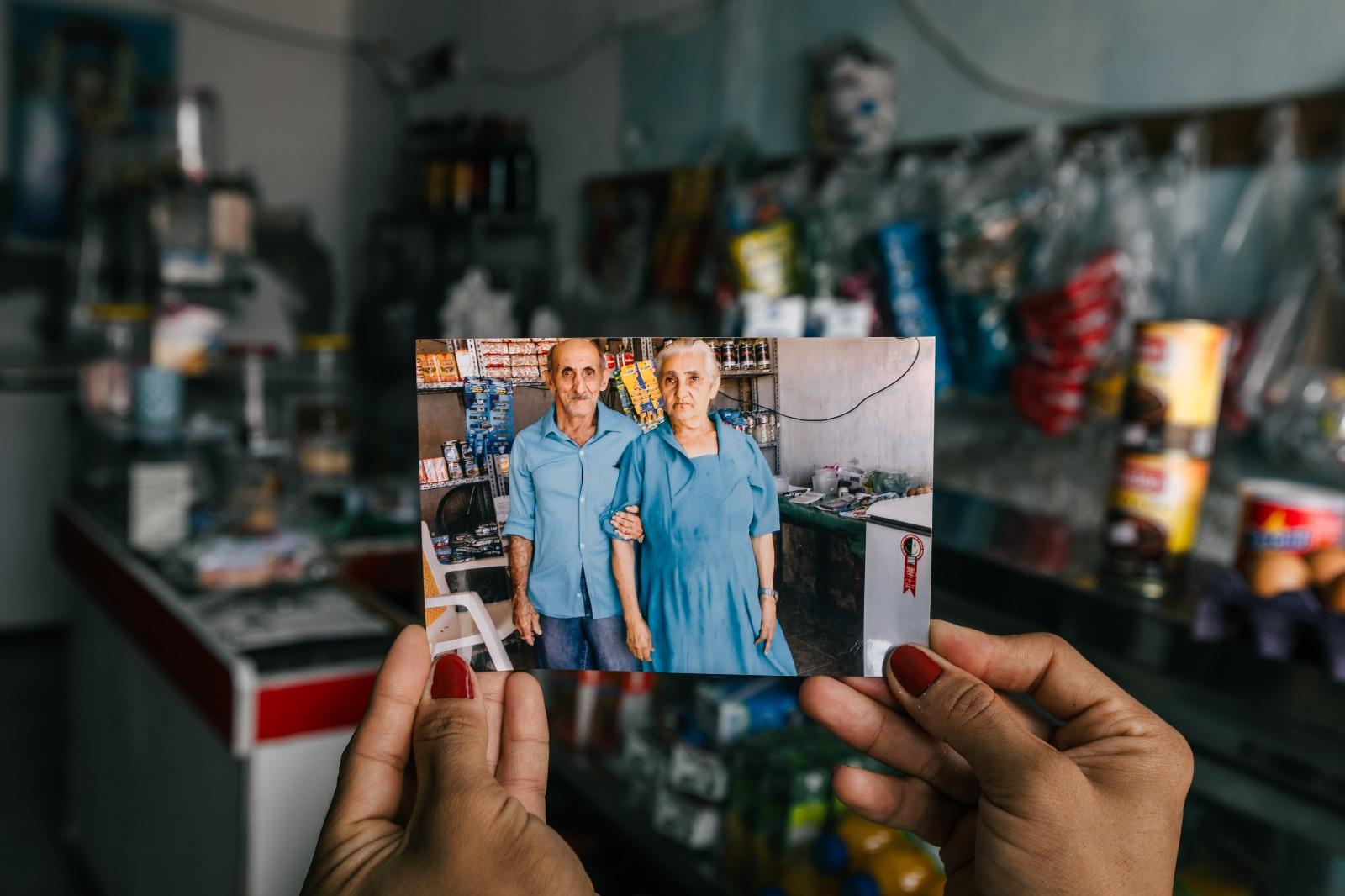Na imagem, uma pessoa segura com ambas as mãos uma fotografia antiga e retangular em frente a um cenário levemente fora de foco. Na foto, um casal de idosos, Seu Francisco e Dona Luzia, aparece em pé dentro de uma pequena mercearia. Ambos vestem roupas azuis e olham para a câmera; ela segura o braço dele delicadamente. Ao fundo, fora de foco, vê-se o interior real da mesma mercearia, com prateleiras repletas de produtos e um balcão, criando uma sobreposição entre o passado registrado na foto e o presente do ambiente.