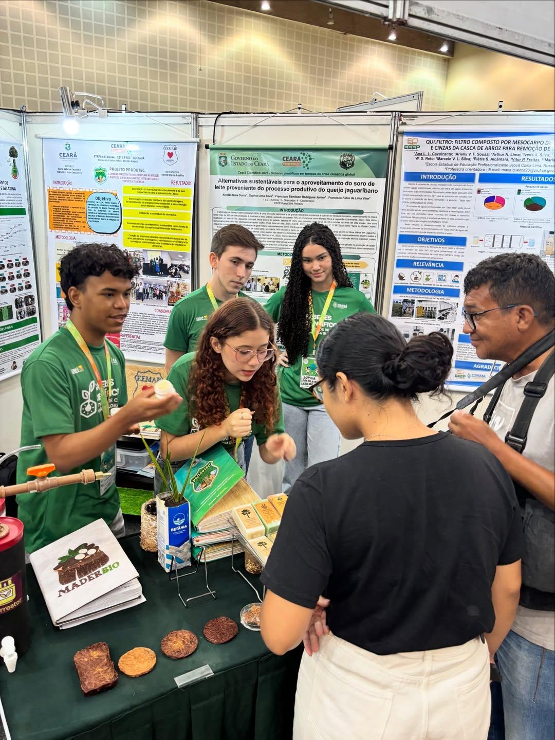 Jovens estudantes em camisetas verdes apresentando produtos alimentícios biológicos e sustentáveis em um estande da feira Ceará Científico.