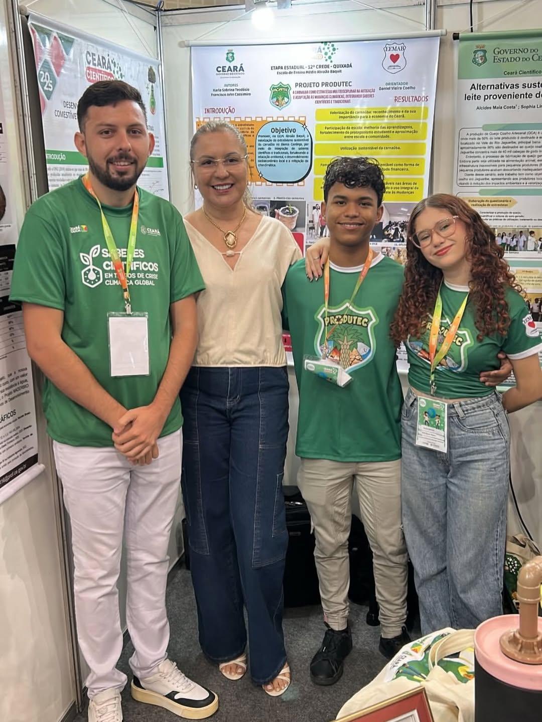 Quatro pessoas, incluindo dois jovens e dois adultos, sorrindo em um estande de exposição no Ceará Científico com pôsteres científicos ao fundo. Dois deles vestem camisetas verdes com o logo Sementes Científicas.