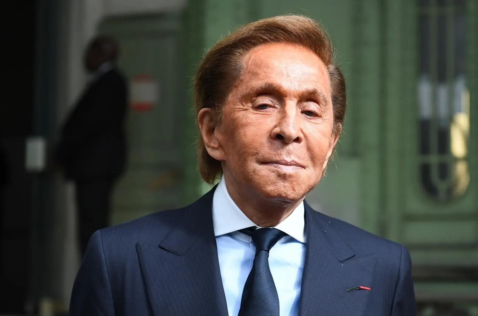 Valentino era um homem idoso, de pele bronzeada e cabelo castanho liso penteado para trás. Na foto, ele usa terno, camisa social e gravata.