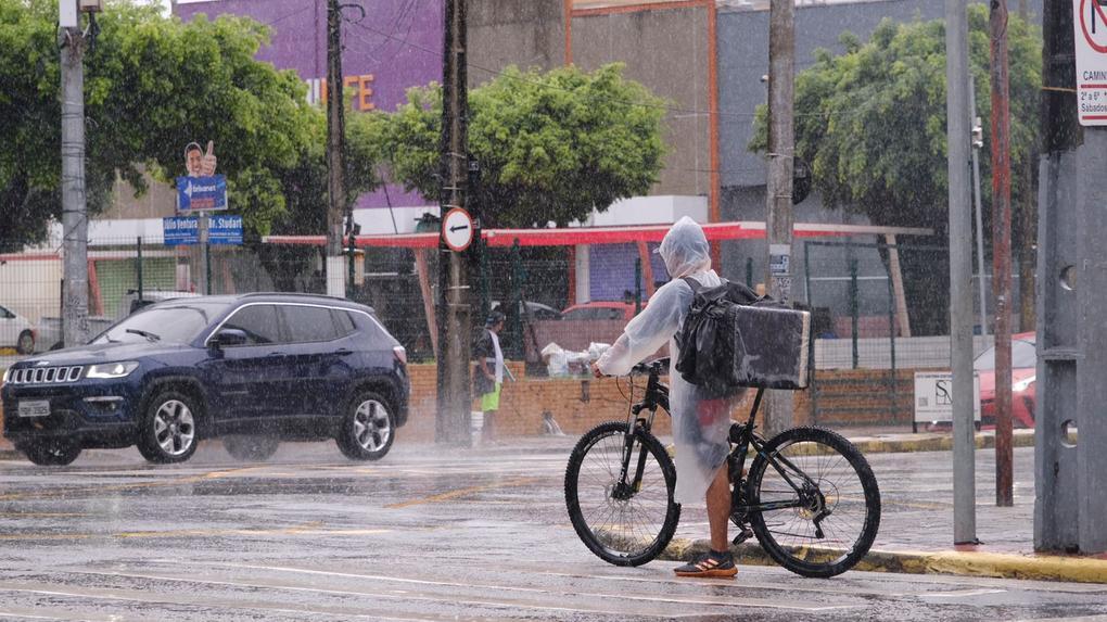 Entregador de bicicleta com capa de chuva em rua de Fortaleza durante temporal. Ao fundo, carros trafegam sob chuva forte e asfalto molhado em cenário urbano.