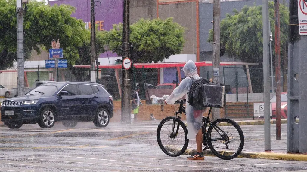 Entregador de bicicleta com capa de chuva em rua de Fortaleza durante temporal. Ao fundo, carros trafegam sob chuva forte e asfalto molhado em cenário urbano.