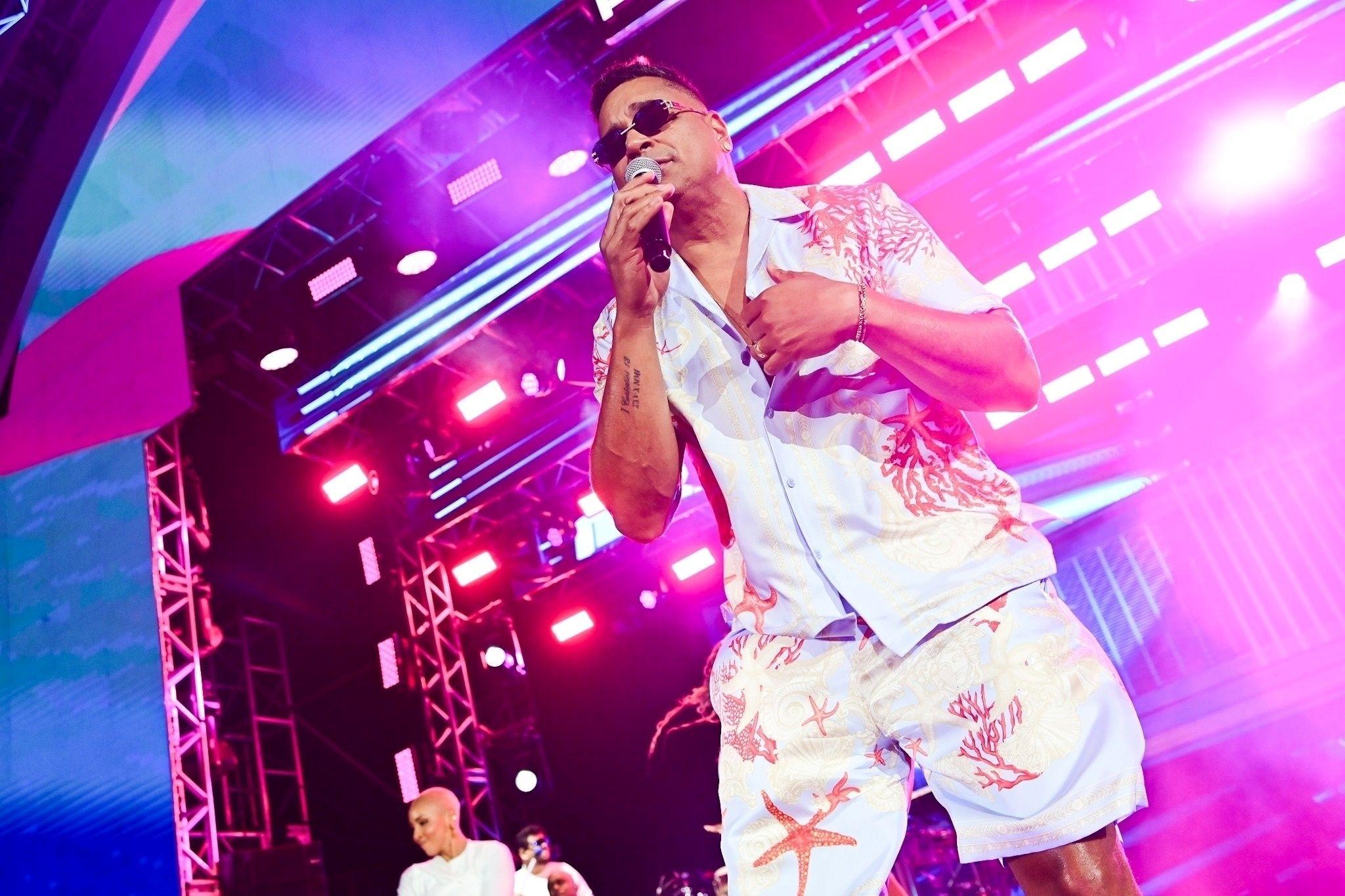 Cantor Xanddy Harmonia com óculos escuros e roupa estampada canta em palco iluminado com luzes roxas e rosas.