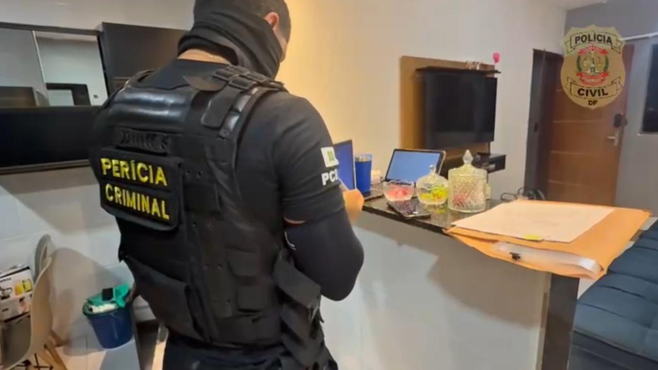 Homem branco fardado com roupa preta da Polícia Criminal e colete, de costas e em uma casa. Na frente dele, há uma bancada com itens diversos e uma televisão na parede.