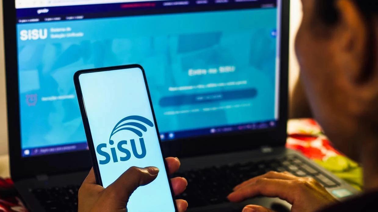 Pessoa utilizando um smartphone com o logo do SISU e um notebook com a página do sistema SISU aberta.