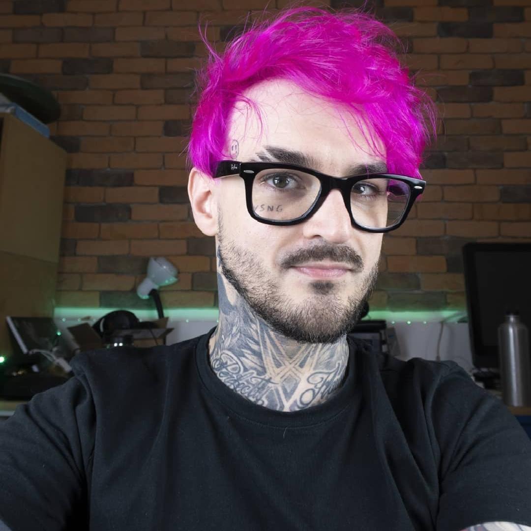PC Siqueira vestindo camisa preta, óculos de armação escura e cabelos na cor roxa.