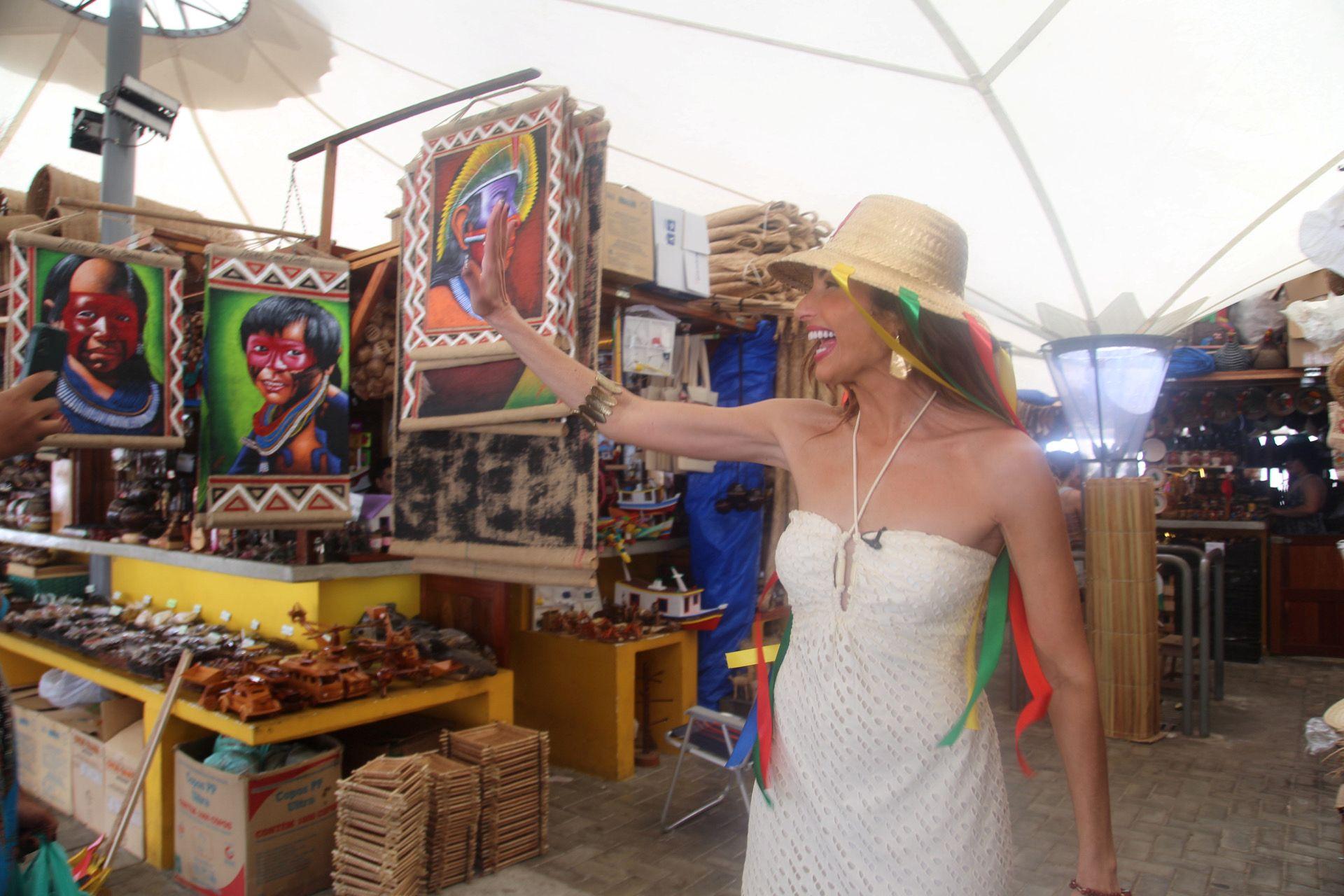 Cena de mercado ao ar livre com Patricia Poeta sorridente em destaque, com obras de arte e produtos artesanais ao fundo.