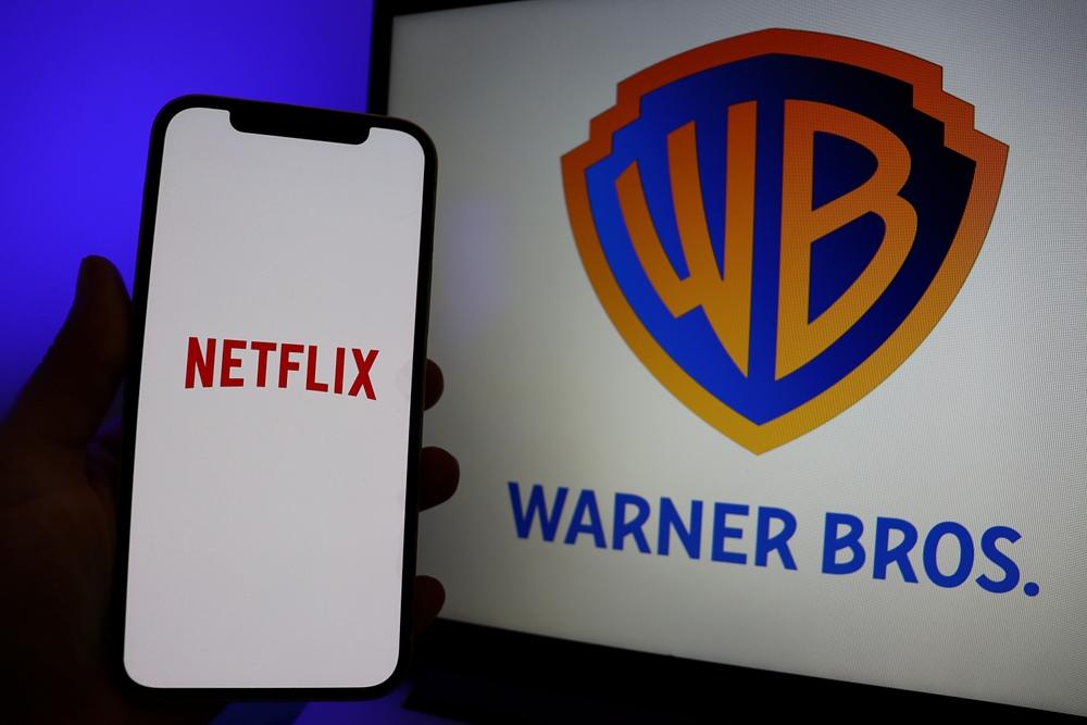 Um smartphone exibindo o logotipo da Netflix em sua tela é segurado diante de um monitor que mostra o logotipo da Warner Bros., com as iniciais “WB” em um escudo colorido e o texto “Warner Bros.” abaixo.