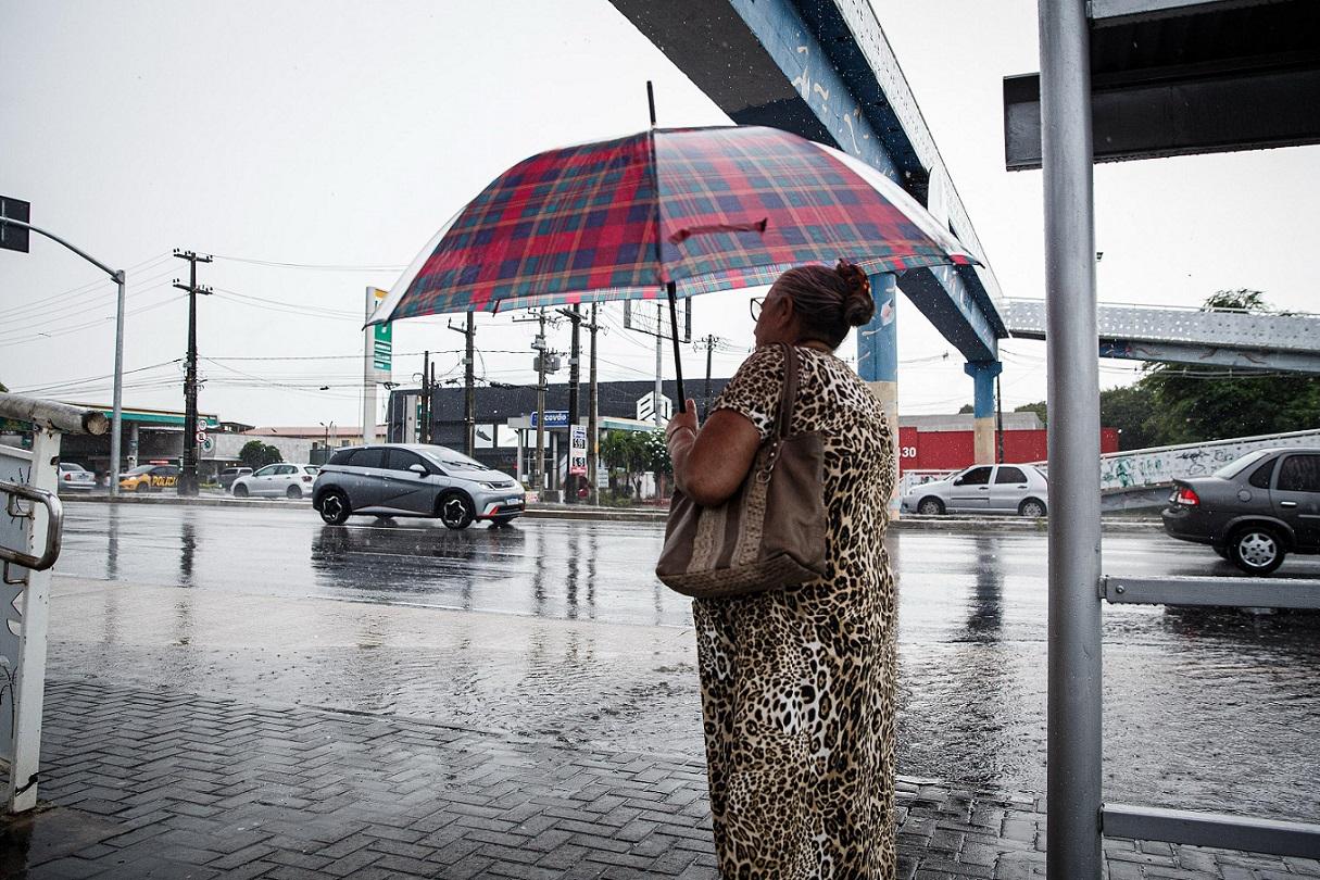 Mulher com vestido de oncinha e guarda-chuva xadrez em uma calçada molhada, observando o tráfego urbano sob uma forte chuva em Fortaleza.