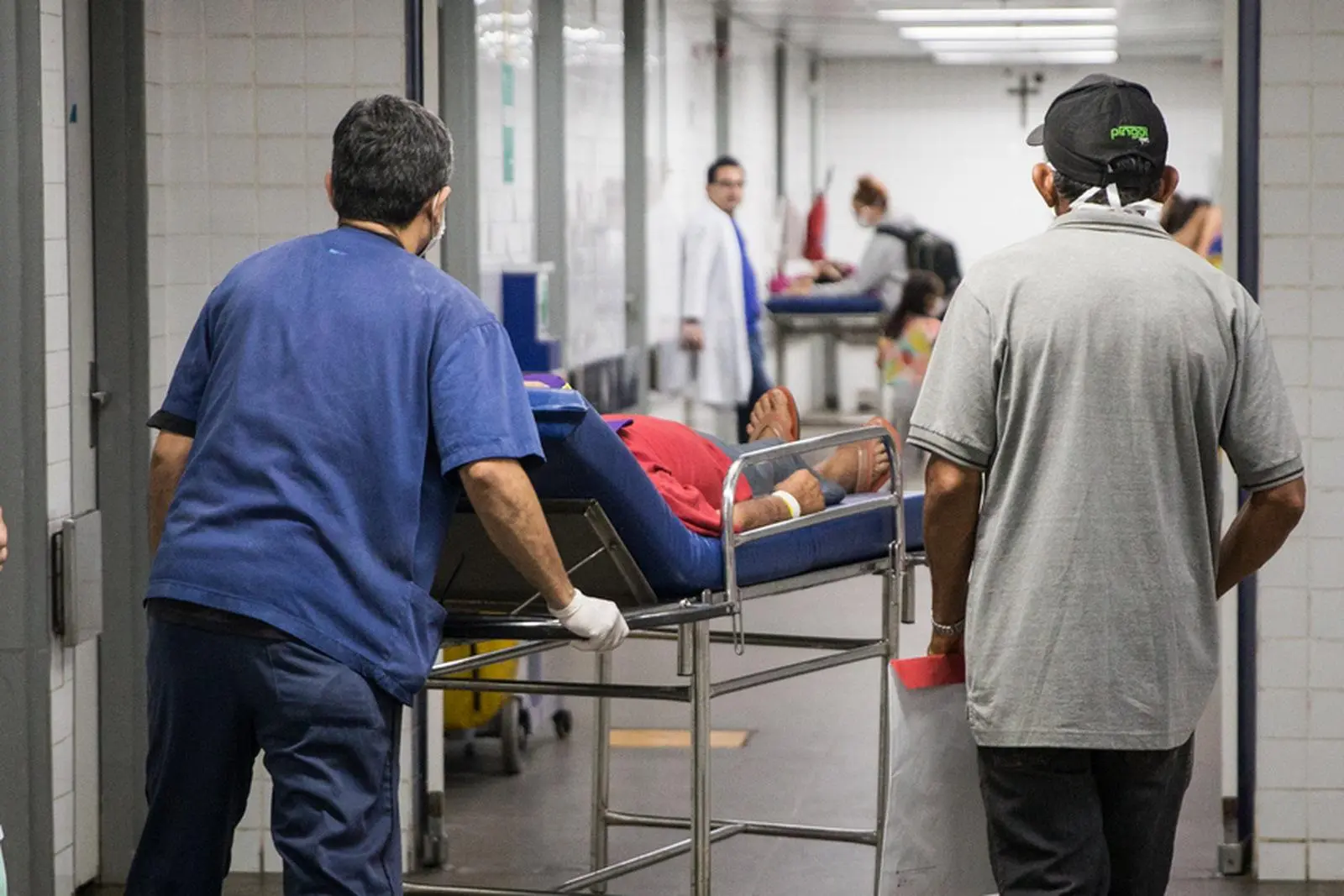Profissionais de saúde transportando paciente em maca por um corredor movimentado de hospital.