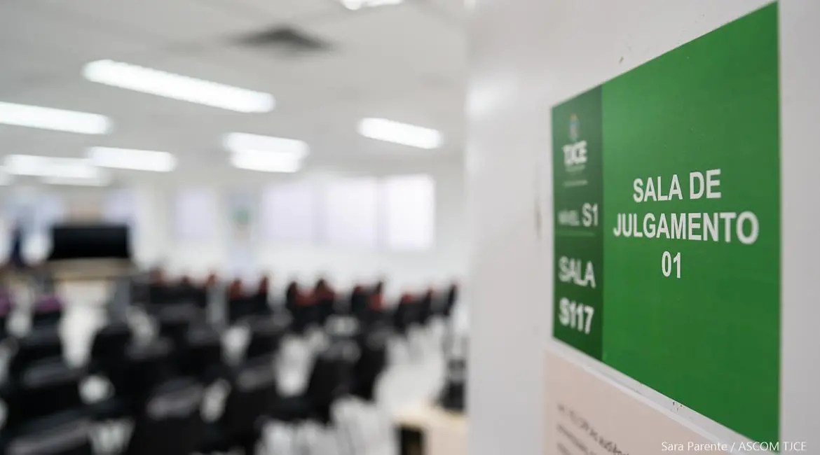 Placa verde sinalizando a sala de julgamento 01 do TJCE, com um auditório ou sala de audiências vazia desfocada ao fundo.