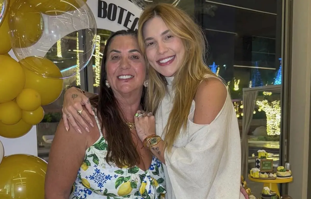 Margareth Serrão ao lado de Virginia. As duas vestem roupas claras e estão sorrindo, posando na frente de balões amarelos.