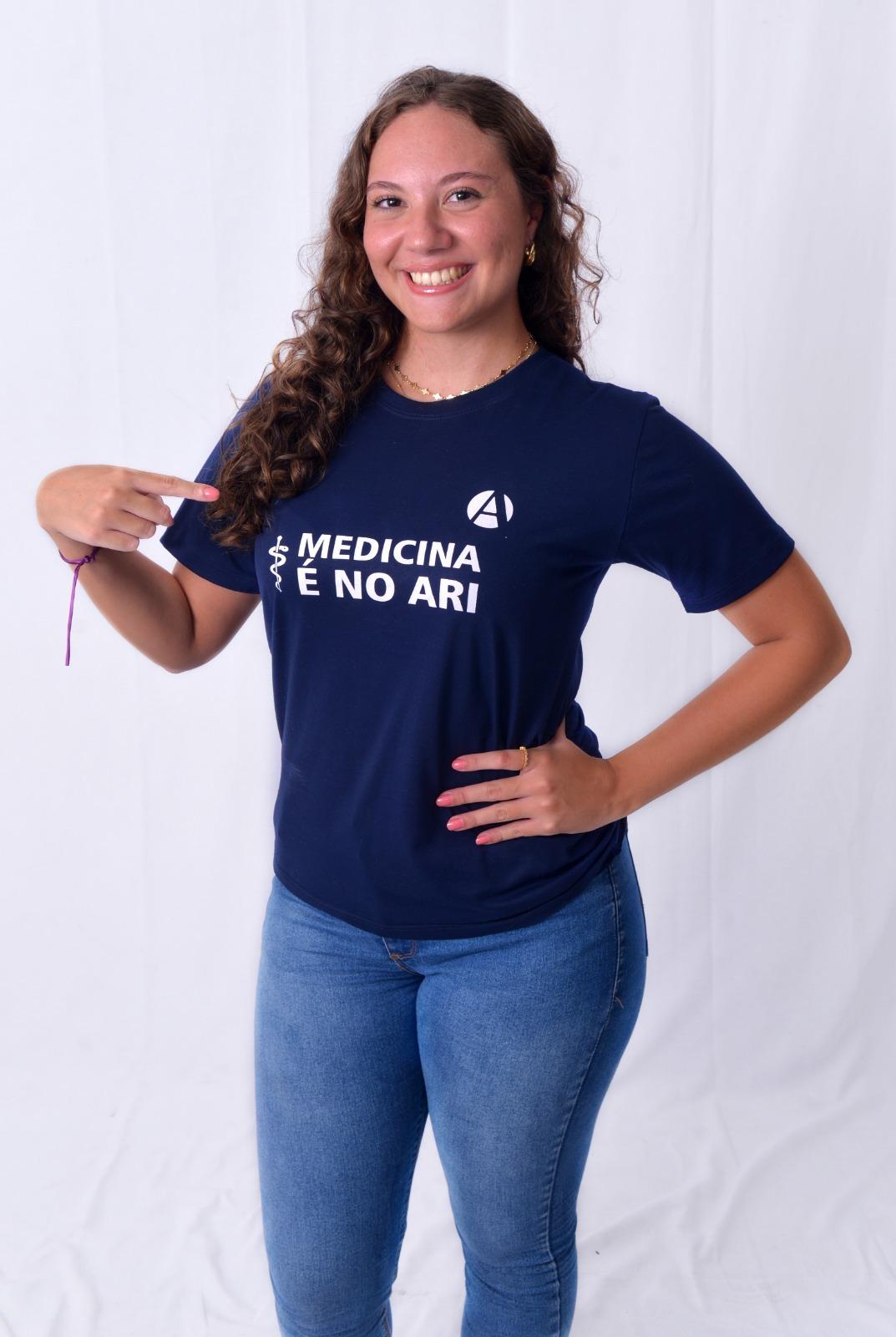 Jovem mulher sorridente com cabelos cacheados castanhos claros, vestindo camiseta com palavra Medicina e apontando para ela.