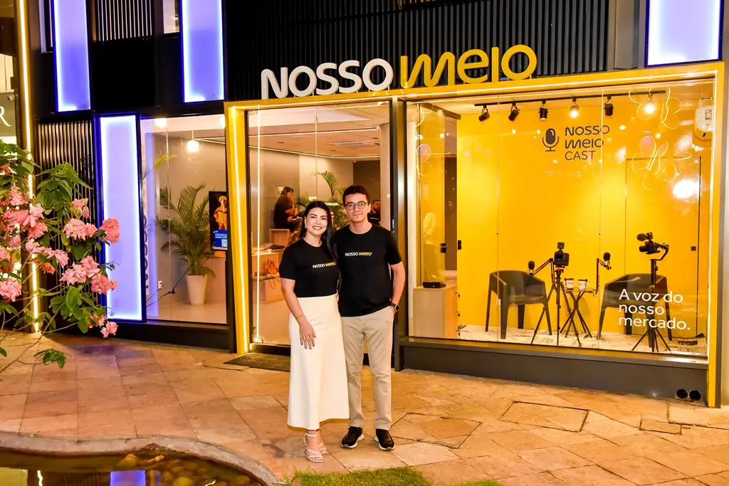 Duas pessoas posam sorrindo em frente ao estúdio do Nosso Meio Cast, com fachada amarela iluminada. Ao fundo, através do vidro, é possível ver o espaço de gravação com cadeiras, microfones, câmeras. A cena é noturna, em um ambiente moderno, com iluminação decorativa e elementos paisagísticos ao redor.