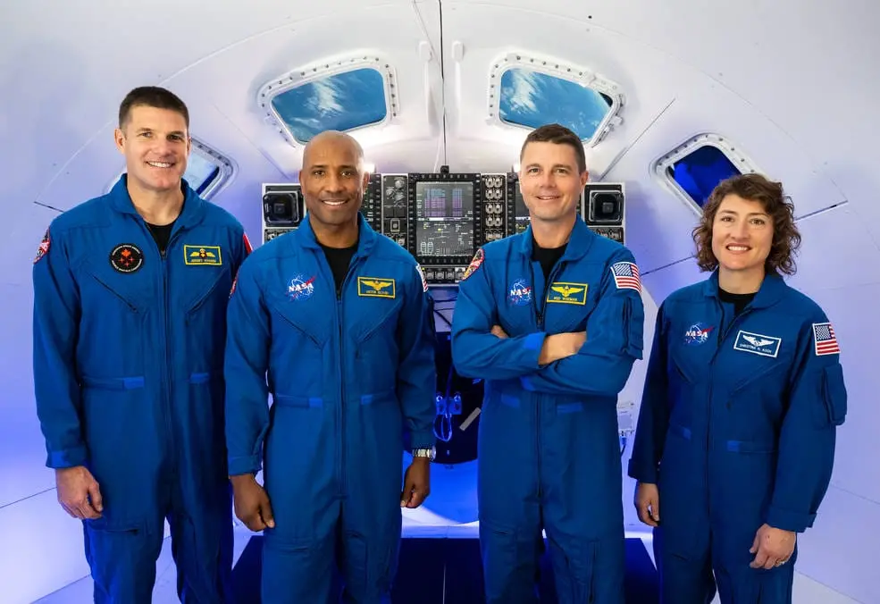 Quatro astronautas vestindo macacões azuis da NASA posam em pé dentro de uma cápsula espacial, com painéis de controle e janelas ao fundo.