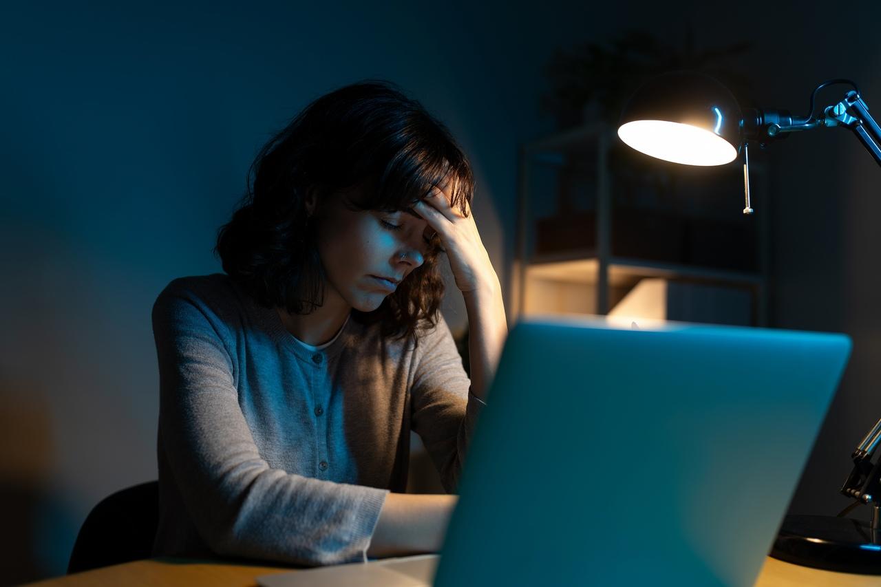 Mulher sentada à mesa à noite, com a mão na testa e expressão de cansaço, trabalhando em um notebook sob a luz de um abajur, representando fadiga e queda de produtividade no trabalho.