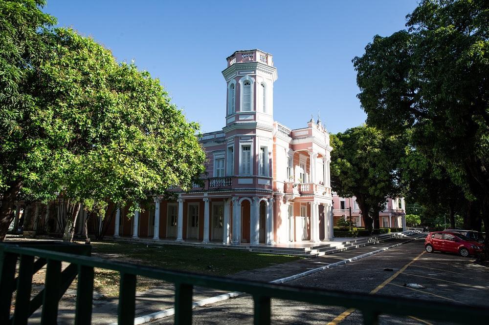 Reitoria da UFC, um edifício histórico rosado e branco com uma torre proeminente, cercado por árvores frondosas e vegetação luxuriante em um dia ensolarado.