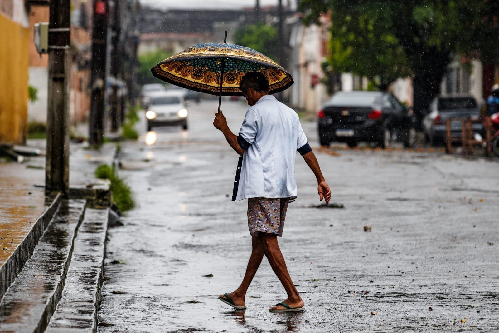 Homem andando por uma rua molhada sob chuva forte, segurando um guarda-chuva estampado para se proteger.